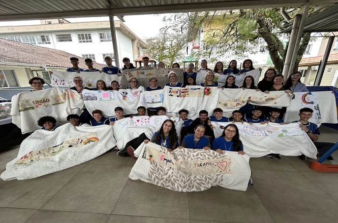 Foto horizontal: grupo de adultos e jovens posa exibindo mantas brancas decoradas com desenhos, palavras e corações.