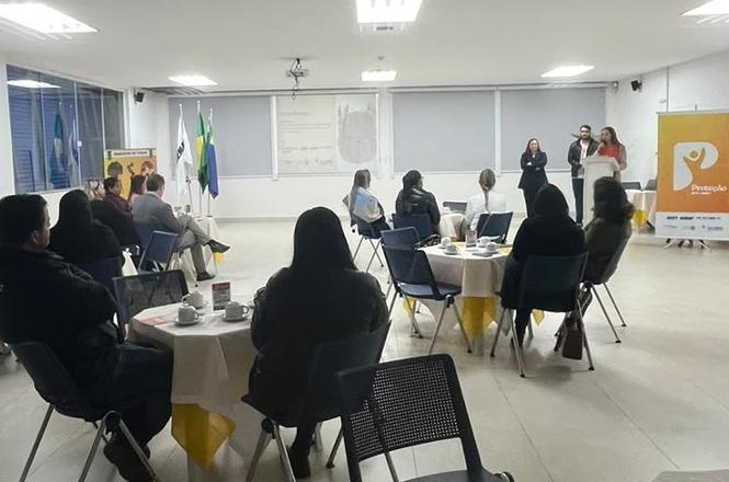 Foto horizontal: participantes sentados em mesas acompanham palestra em sala, com três pessoas à frente.