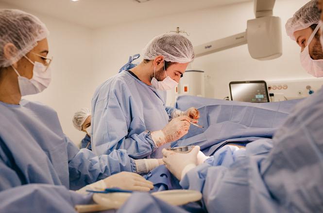 Foto horizontal: equipe médica com roupas azuis, máscaras e toucas realiza procedimento cirúrgico em centro hospitalar.