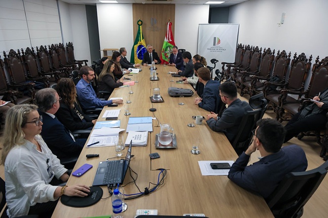 Foto horizontal: autoridades e servidores participam de reunião em mesa ampla, com bandeiras do Brasil e de SC ao fundo.