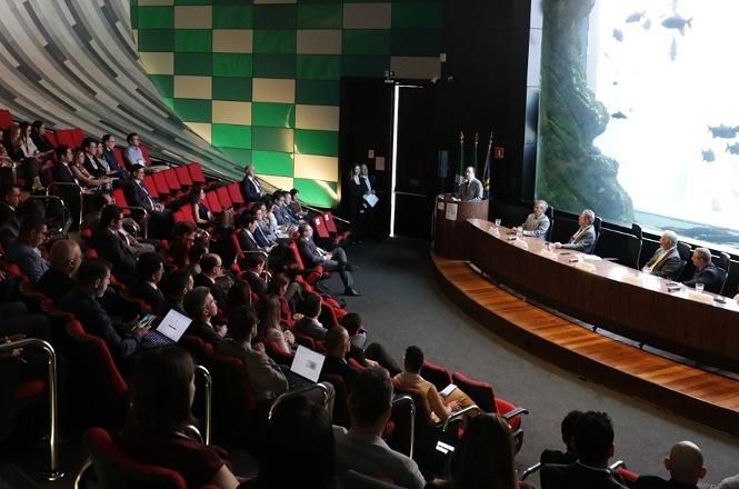 Foto horizontal: público ocupa auditório com poltronas vermelhas durante solenidade, com autoridades à mesa.