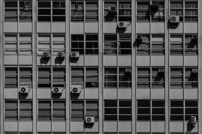 Foto horizontal em preto e branco: fachada de edifício com fileiras de janelas e aparelhos de ar-condicionado.
