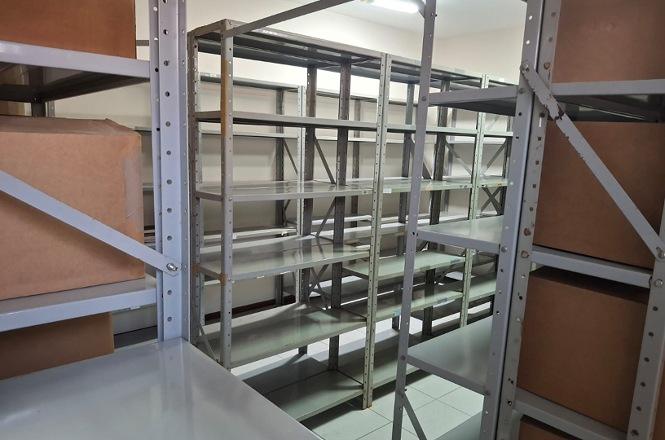 Foto horizontal: arquivo com estantes metálicas, prateleiras vazias e caixas de papelão armazenadas nas laterais.