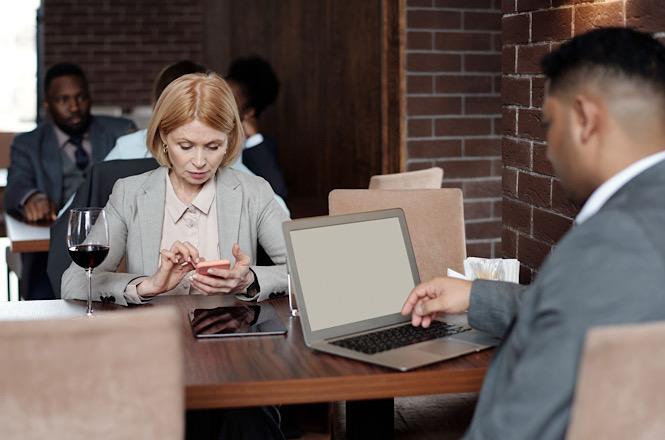 Foto horizontal: mulher de blazer cinza usa celular diante de homem com notebook aberto, em mesa de restaurante.