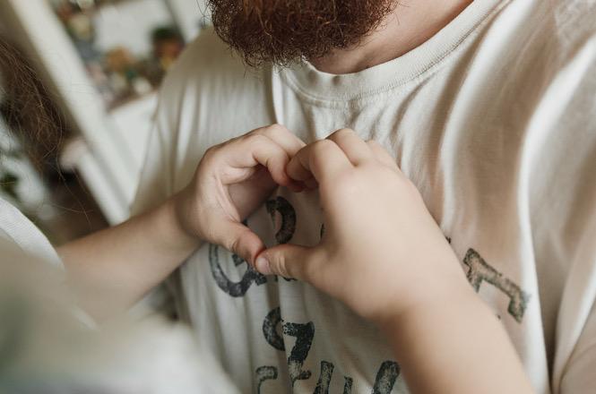 Foto horizontal: criança forma coração com as mãos sobre o peito de homem barbado com camiseta branca.