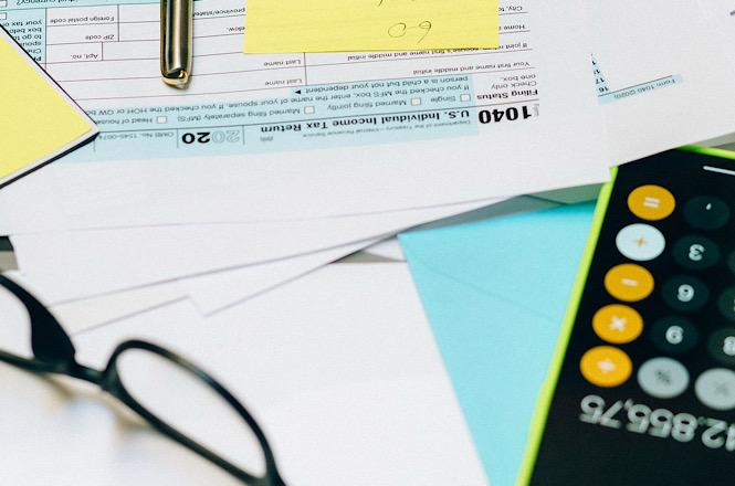Foto horizontal: documentos fiscais, calculadora, óculos e papéis coloridos estão dispostos sobre uma mesa.