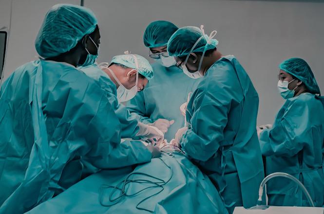 Foto horizontal: equipe médica realiza procedimento cirúrgico em paciente, com profissionais usando trajes verdes, máscaras e toucas.