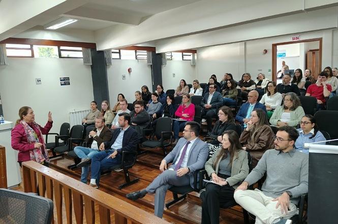 Foto horizontal: desembargadora Hildemar Meneguzzi de Carvalho, de blusa rosa, mostra o sinal vermelho na palma da mão diante do público.