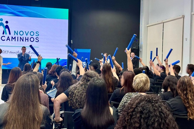 Foto horizontal: participantes do programa Novos Caminhos erguem bastões azuis durante atividade coletiva em auditório.