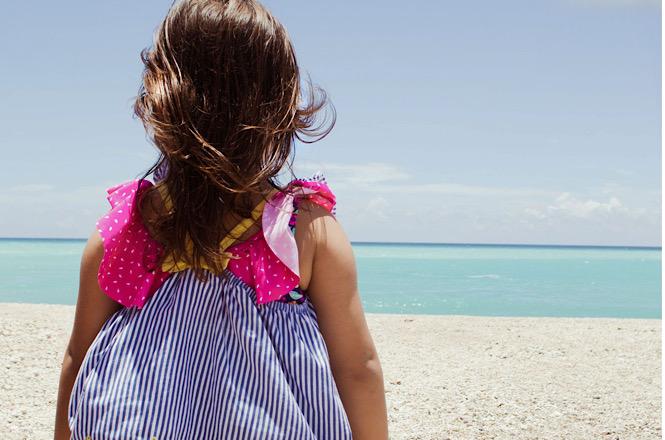 Foto horizontal: criança de vestido rosa e azul observa o mar de águas claras, de costas, em praia ensolarada.