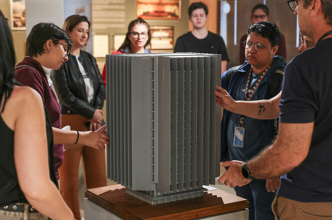 Foto horizontal: visitantes observam e tocam maquete de edifício durante exposição em espaço cultural.