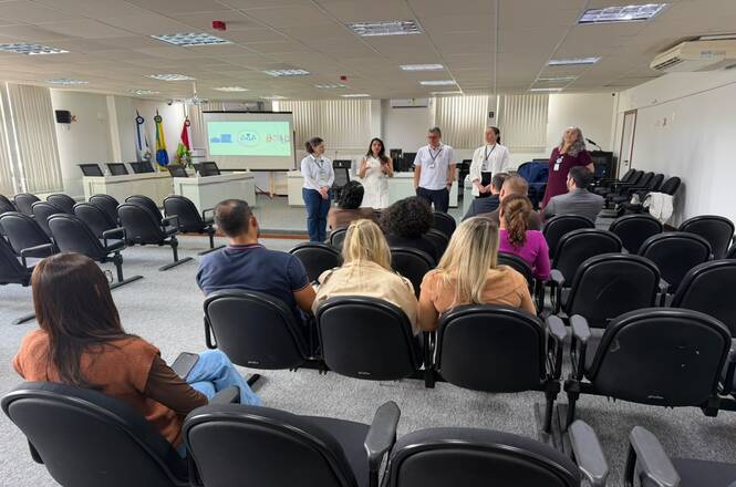 Foto horizontal: participantes acompanham atividade em auditório, enquanto equipe apresenta informações diante do público, com tela de projeção e bandeiras ao fundo.