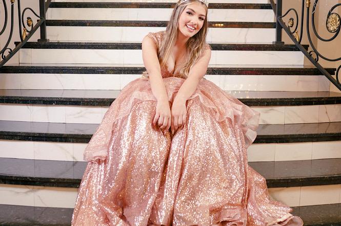 Foto horizontal: jovem posa sorridente em escadaria, usando vestido longo rosa com brilho e tiara, em traje de festa.