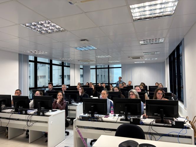Fotografia de uma sala de informática durante uma capacitação presencial. Cerca de 20 participantes estão sentados em mesas organizadas em fileiras, cada um diante de um computador de mesa. A imagem foi capturada de frente para a turma, mostrando os participantes voltados para a área do instrutor, localizada fora do enquadramento. O ambiente possui paredes claras, iluminação por painéis no teto e grandes janelas nas laterais, que permitem a entrada de luz natural e revelam edifícios ao redor. Sobre as mesas, há monitores, teclados, mouses, cabos e alguns objetos pessoais, como garrafas de água. A disposição do espaço evidencia uma atividade de treinamento voltada ao uso de recursos tecnológicos.