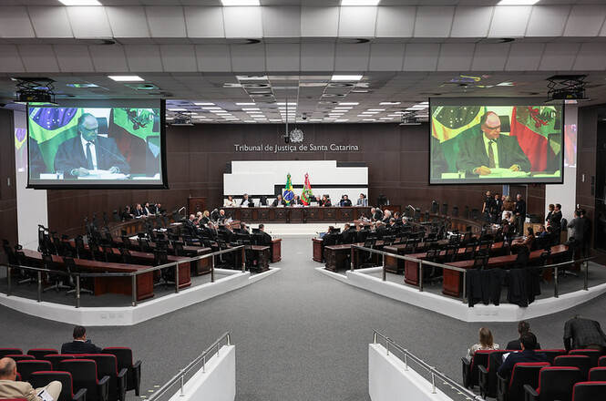 Foto horizontal: sessão no Tribunal Pleno do TJSC reúne magistrados no plenário, com desembargador em destaque nos telões e bandeiras ao fundo.