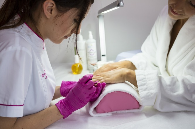 Foto horizontal: profissional realiza procedimento de manicure nas unhas de cliente, com luvas rosas e materiais de cuidado sobre a bancada.