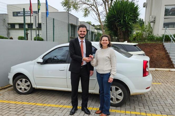 Foto horizontal: homem de terno entrega a chave de um veículo branco a uma mulher, em área externa de unidade do Poder Judiciário.