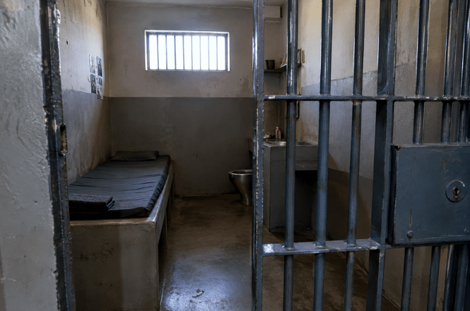 Foto horizontal: interior de cela prisional vazia, com cama de concreto e colchão escuro à esquerda, vaso sanitário metálico ao fundo, pequena janela com grades e porta de ferro gradeada em primeiro plano.