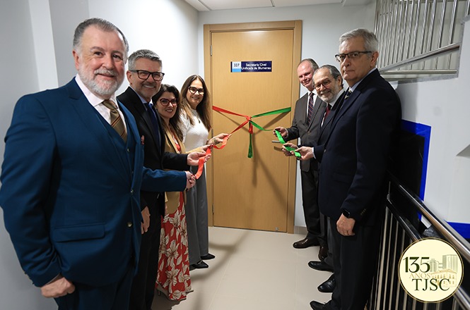 Foto horizontal: autoridades e servidores do TJSC participam de cerimônia de inauguração de sala, com corte simbólico de fitas nas cores verde e vermelha, diante da porta de entrada, em corredor institucional.