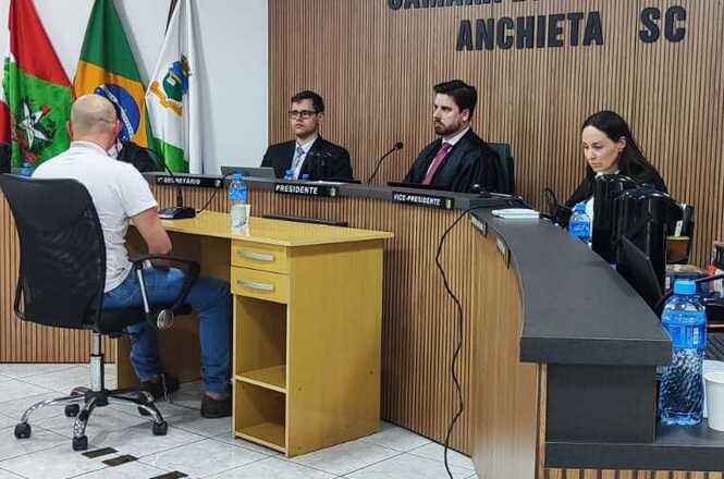 Foto horizontal: homem de camiseta branca, sentado de costas, participa de audiência na Câmara Municipal de Anchieta, diante de dois homens de terno identificados como presidente e vice-presidente e de uma mulher que acompanha documentos à mesa. Ao fundo, bandeiras de Santa Catarina, do Brasil e do município.