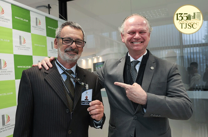 Foto horizontal: oficial de justiça Paulo Roberto Costa, à esquerda, exibe crachá de identificação ao lado do desembargador Rubens Schulz, que sorri e aponta para o servidor, diante de painel institucional verde e branco do TJSC.
