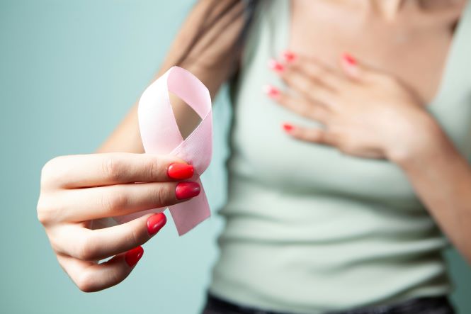 Imagem em close mostra uma pessoa segurando um laço rosa com a mão estendida em direção à câmera. Ao fundo, desfocado, aparece o tronco da pessoa com uma das mãos apoiada sobre o peito. A fotografia utiliza tons suaves e remete à campanha Outubro Rosa, de conscientização sobre a prevenção e o diagnóstico precoce do câncer de mama.