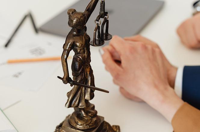 Foto horizontal: estatueta de bronze da Justiça, segurando balança e espada, aparece em primeiro plano, com mãos de pessoa de terno apoiadas sobre mesa com documentos ao fundo.