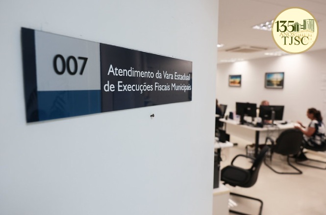 Foto horizontal: placa identifica a sala 007, destinada ao atendimento da Vara Estadual de Execuções Fiscais Municipais, com servidores trabalhando em computadores ao fundo.