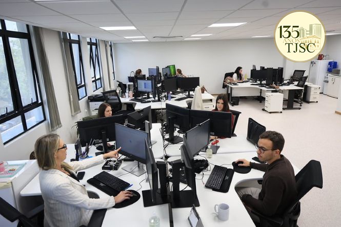 Foto horizontal: servidores do TJSC trabalham em escritório compartilhado, diante de computadores com múltiplos monitores, mesas brancas e janelas amplas.