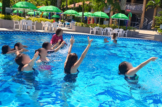 Foto horizontal: grupo de mulheres participa de atividade aquática em piscina azul, com braços erguidos, enquanto instrutora orienta os movimentos; ao fundo, mesas, cadeiras e guarda-sóis verdes.