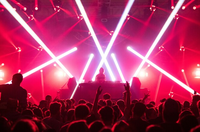 Foto horizontal: DJ se apresenta em palco durante festa de música eletrônica, com feixes de luz branca e vermelha cruzando o ambiente, enquanto o público acompanha a apresentação com os braços erguidos.