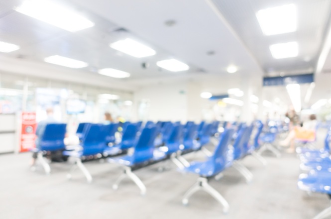 Foto horizontal: sala de espera de ambiente de atendimento ao público, com fileiras de cadeiras azuis, piso cinza e iluminação branca no teto, em imagem desfocada.
