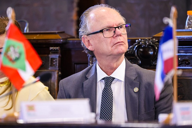 Foto horizontal: presidente do TJSC, desembargador Rubens Schulz, de terno cinza, gravata azul e óculos, aparece sentado durante solenidade institucional, olhando para o lado, com bandeiras de Santa Catarina e de outros estados em primeiro plano.