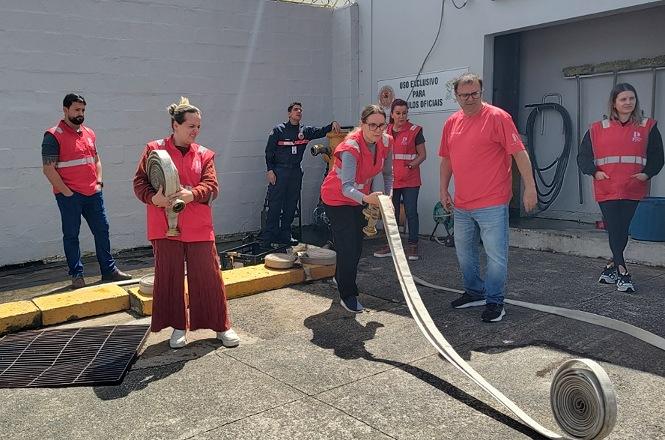 Foto horizontal: servidores e brigadistas, com coletes vermelhos, participam de treinamento de combate a incêndio em área externa, realizando o manuseio e a extensão de mangueiras, sob orientação de profissionais.