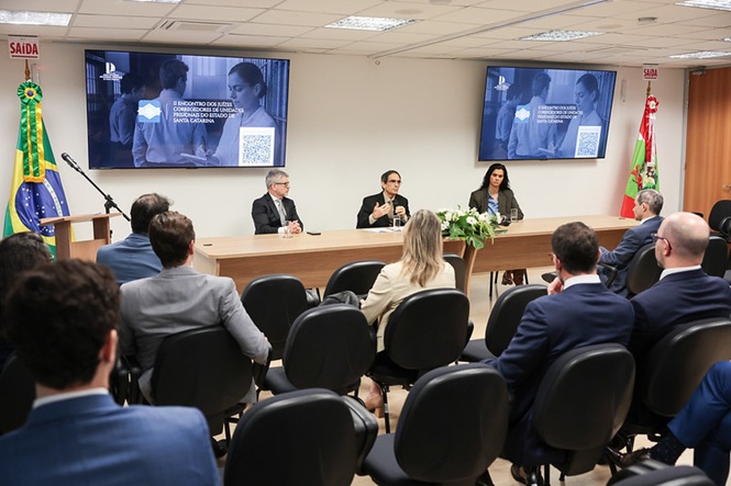 Horizontal: O desembargador Roberto Lucas Pacheco, o corregedor-geral da Justiça, Dinart Francisco Machado, e a juíza Fernanda Pereira Nunes participam de mesa de abertura em encontro institucional, realizado em auditório com telões de apresentação, bandeiras oficiais, arranjo floral e público acomodado em fileiras de cadeiras.