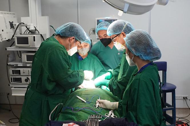 Horizontal: Equipe médica realiza procedimento cirúrgico em centro cirúrgico hospitalar, com profissionais paramentados ao redor da mesa operatória, iluminação focal sobre o campo cirúrgico, equipamentos de monitoramento, aparelhos médicos e mobiliário clínico