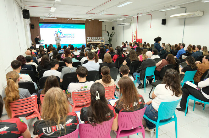 Foto horizontal de auditório lotado durante palestra do programa Novos Caminhos, com participantes sentados em cadeiras coloridas acompanhando apresentação em telão branco, azul e verde.  Forneça seus comentários sobre o BizChat