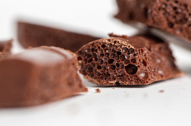 Horizontal: Close-up de pedaços de chocolate aerado sobre superfície clara, com textura porosa em destaque e fundo desfocado que evidencia as bolhas internas do alimento.