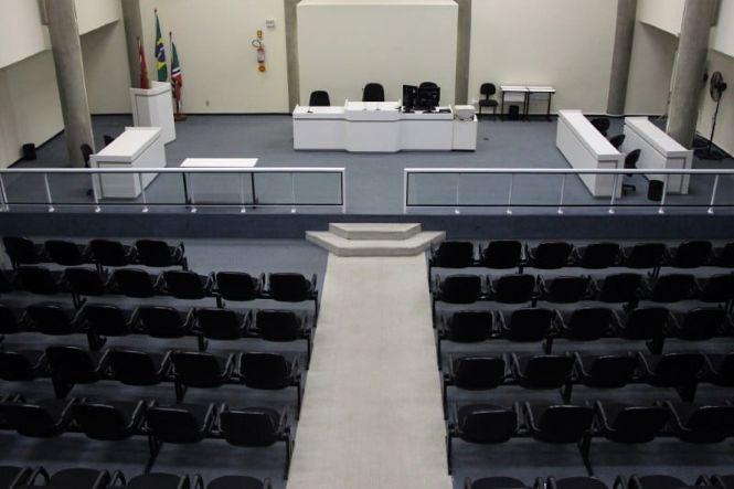 Foto colorida: plenário de audiências vazio, sala judicial interna, estrutura do fórum, exibindo espaço em branco, cinza e preto