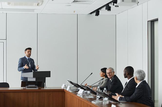 Foto colorida: pessoa discursando em púlpito, sala de reuniões institucional, sessão administrativa, apresentando propostas em branco, cinza e marrom