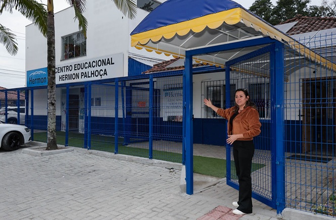 Foto colorida: mulher usando casaco marrom e calça preta diante do Centro Educacional Hermon Palhoça, área externa urbana, apresentando a entrada em azul, branco e marrom.