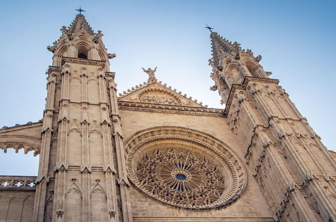 Horizontal: Fachada de catedral em estilo gótico, com duas torres ornamentadas, rosácea central esculpida e céu azul ao fundo.