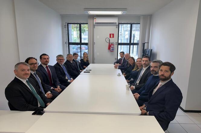 Horizontal: Participantes posam ao redor de mesa de reuniões em sala institucional, organizada em formato retangular. O ambiente possui paredes brancas, janelas com grades, aparelho de ar-condicionado, extintor de incêndio fixado na parede
