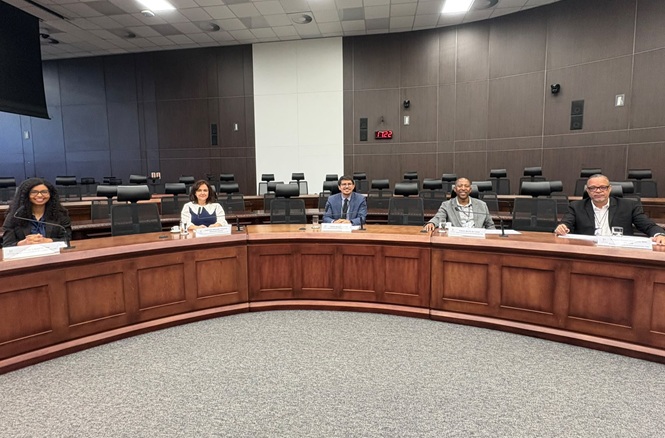 Horizontal: Participantes acompanham reunião em sala de sessões, sentados atrás de bancada semicircular de madeira em ambiente amplo com cadeiras para o plenário