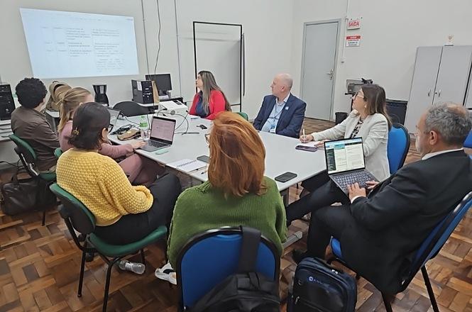 Foto colorida: participantes em reunião à mesa, sala institucional interna, encontro de trabalho, acompanhando apresentação em branco, azul e verde.