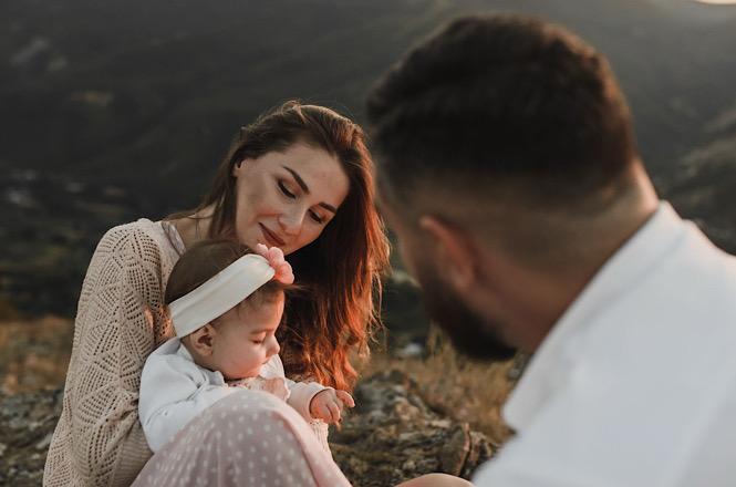 Foto horizontal: mãe segura bebé no colo com pai em primeiro plano, ao ar livre e ao pôr do sol nas montanhas.