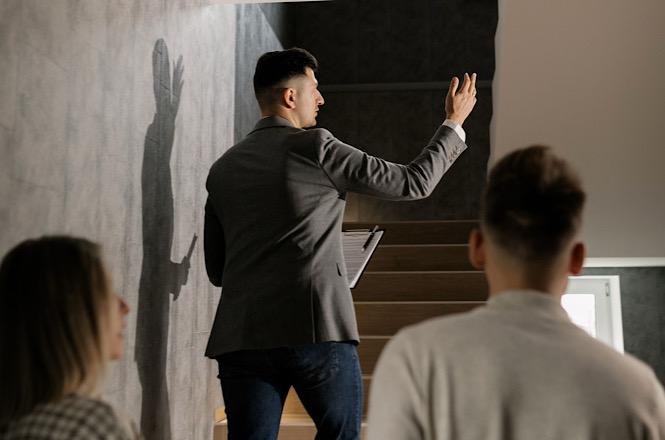 Foto horizontal: homem de blazer gesticula na escada diante de casal, projetando sombra na parede.