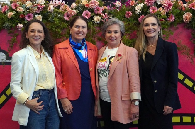 Foto horizontal: a desembargadora Hildemar Meneguzzi (de blazer vermelho e lenço azul) posa com outras três mulheres elegantes em frente a um painel cor-de-rosa decorado com flores naturais