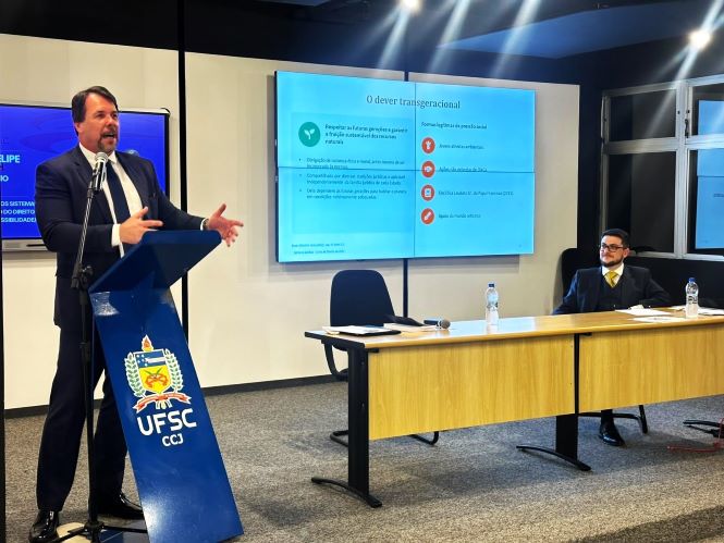 Em ambiente de palestra, desembargador Luiz Felipe Schuch, de terno escuro fala ao microfone em um púlpito azul com o brasão e a sigla da UFSC, gesticulando durante a apresentação. Ao fundo, uma tela exibe o slide “O dever transgeracional”. À direita, o servidor Marlio Aguiar, de terno, está sentada à mesa, acompanhando a exposição.