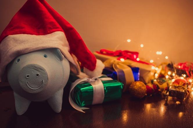 Foto horizontal: mealheiro em forma de porquinho azul com barrete de Pai Natal em primeiro plano, ao lado de pequenos presentes embrulhados e luzes natalícias desfocadas ao fundo.
